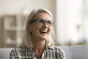 Smiling older woman with straight, shoulder-length gray hair and glasses, wearing a plaid shirt, sits indoors on a couch. The background is softly blurred, featuring neutral tones and sunlight.