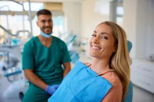 A smiling woman sits in a dental chair wearing a blue bib, looking at the camera. A male dentist in green scrubs with gloves sits behind her in a bright, modern dental office.