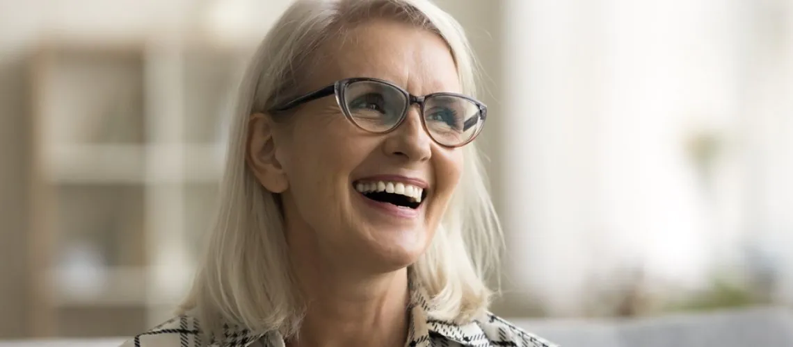 Smiling older woman with straight, shoulder-length gray hair and glasses, wearing a plaid shirt, sits indoors on a couch. The background is softly blurred, featuring neutral tones and sunlight.