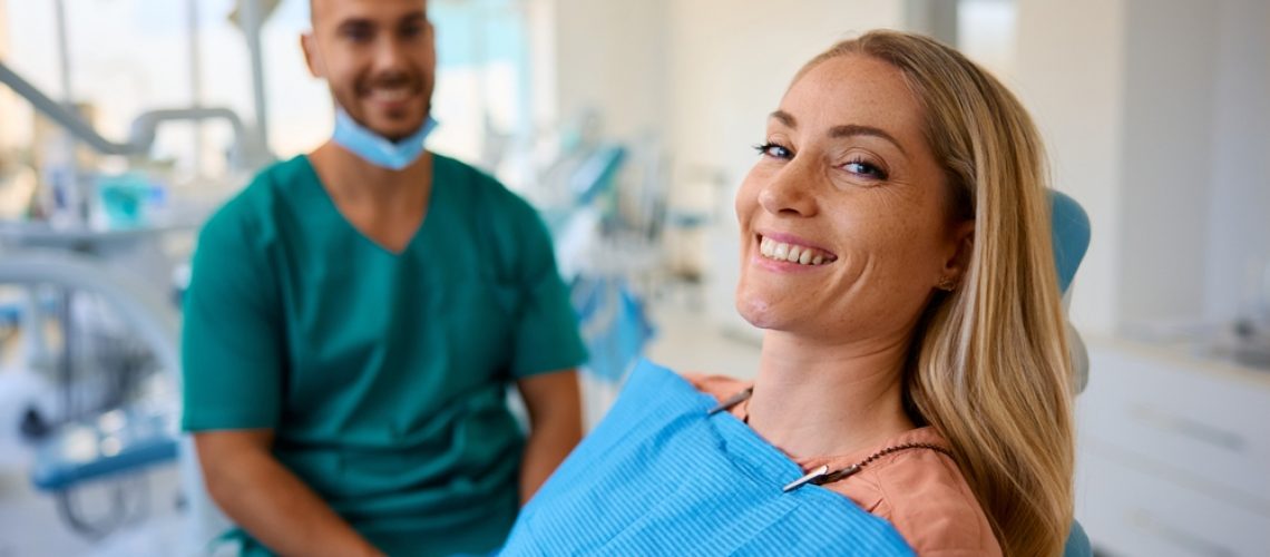 A smiling woman sits in a dental chair wearing a blue bib, looking at the camera. A male dentist in green scrubs with gloves sits behind her in a bright, modern dental office.